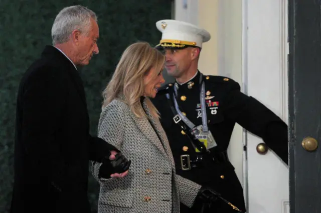 Kennedy in all black holds the hand of his wife in a black and white blazer as a military person in uniform opens a door for them