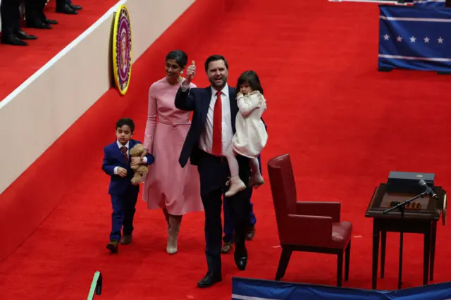 Usha and JD Vance walk on a red carpet while holding their young children