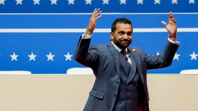 Kash Patel with his arms raised on a stage with a blue banner with white stars in the background