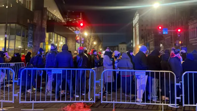 A huge crowd of people who are queuing, held in line by metal barriers. It's dark outside and people are wrapped up warm