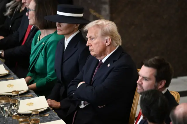 Donald Trump, with his arms folded, sitting at a table set for lunch with Melania Trump seated on his left and JD Vance on his right