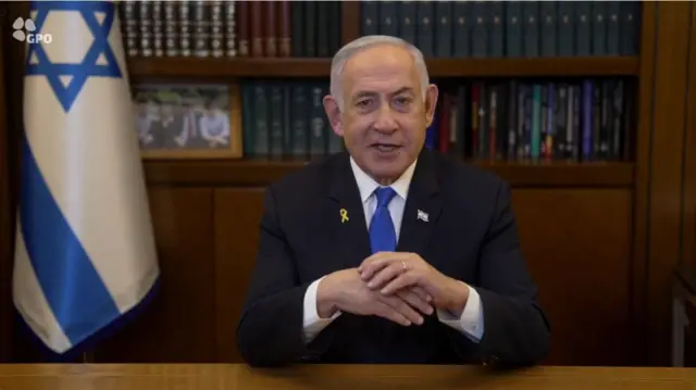 Benjamin Netanyahu delivers congratulatory speech for the inauguration of Donald Trump. He's sitting down at a wooden desk in a black suit, leaning on his elbows as his left hand rests on top of his right one. There's a wooden bookcase in the background and a Star of David flag to his right