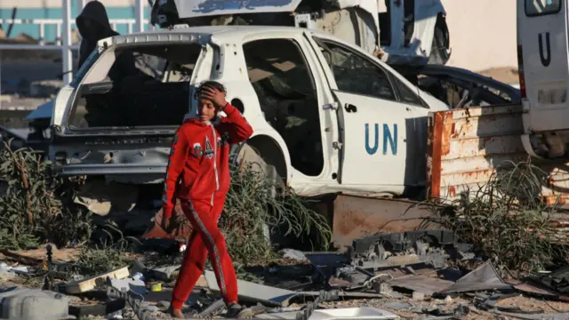 A girl wearing a red tracksuit walks past a destroyed white vehicle with 'UN' written on the door.