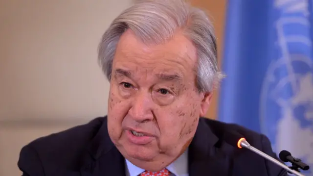 Close up shot of Antonio Guterres delivering a speech sitting down. He's in a black suit, light blue shirt and red tie, two mics in front of him and a UN flag (light blue background and UN logo) blurred