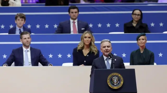 Steve Witkoff speaking on stage at the inauguration parade, with members of the Trump family sat behind him