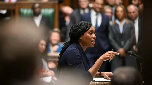 Kemi Badenoch speaks in the House of Commons holding a pen and pointing forward with MPs blurred out in the background.