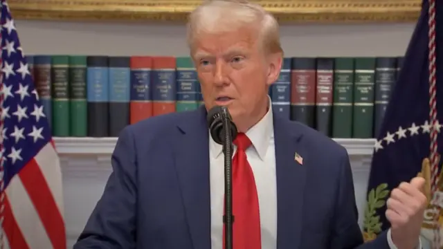 Trump, in a blue suit and red tie, gestures beside him with his thumb to point at the people offscreen