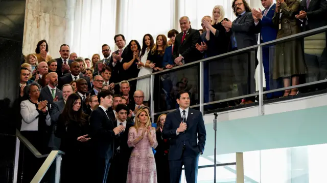 Rubio in dark suit with microphone and hand in pocket stands on stairs with crowd behind him