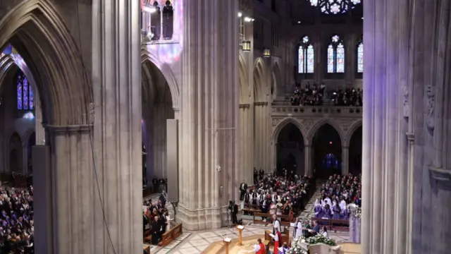 A view from the above balconies shows a general view of the prayer service at Washington National Cathedral. Attendees sit on chairs in rows with the cathedral's reverend sat in the centre of the building.