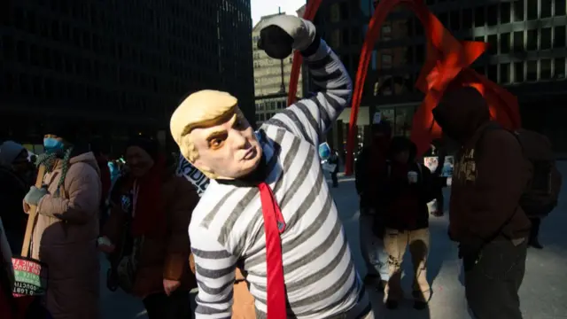 A person with a Trump mask over his head, wearing a striped long sleeved shirt and red tie. He is raising his bicep.