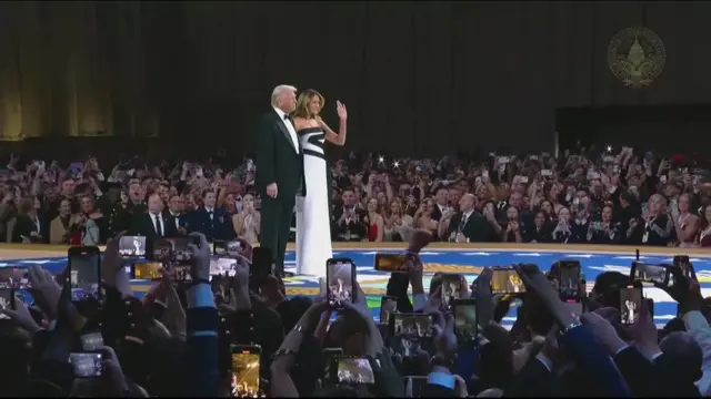 Trump and Melania stand on stage at the ball. Melania, in a white dress with black detailing, waves at the crowd