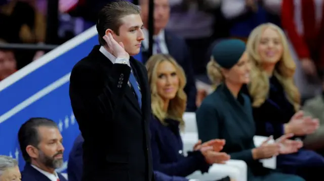 Barron Trump in a suit and blue tie gesturing to crowd.