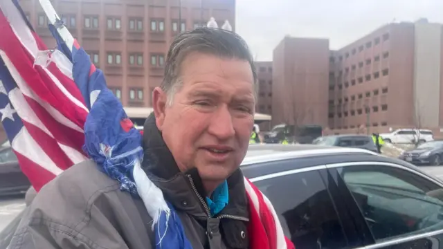 Ben Pollock holds a US flag outside the DC central jail. He has short, dark hair and is wearing a black coat.