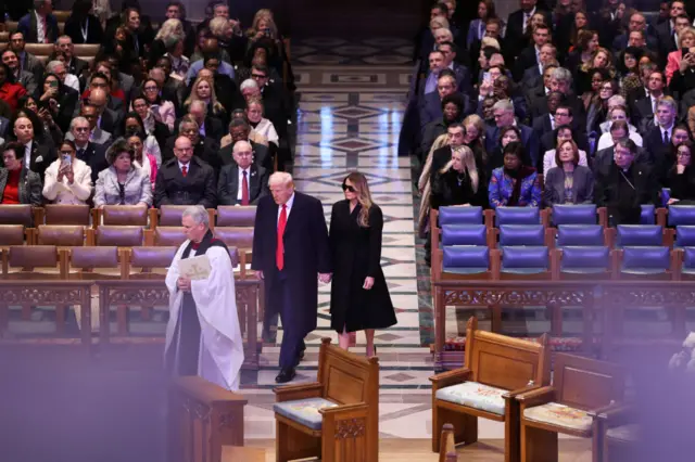 President Donald Trump and First Lady Melania Trump walk to the front row of seats in Washington's National Cathedral. Other attendees sit behind them.