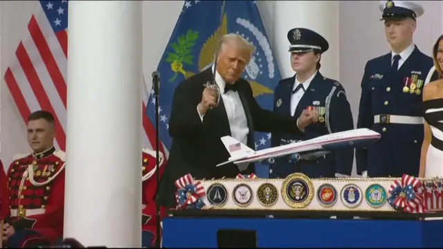 Trump in a suit dances with a thin rapier-like sword held aloft in front of a large cake shaped like his airplane