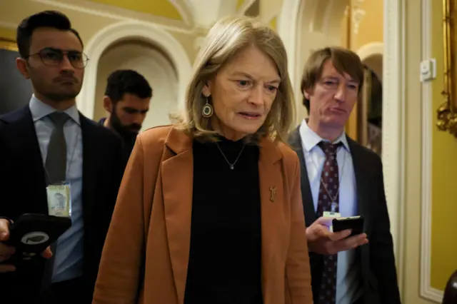 Alaska state Senator Lisa Murkowski walking inside the US Capitol flanked by colleagues. She has short hair and wears an orange jacket.