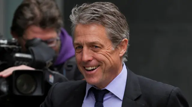 Hugh Grant walking past a cameraman with a camera outside the High Court in 2023. He is smiling and wearing a black blazer, light purple shirt, and dark navy tie. He has blue eyes and greying brown hair.