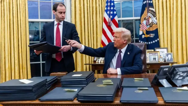 Trump sits at a desk in the Oval Office in the White House in a suit and points. His desk is covered in various blue books.