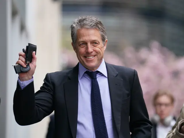 Hugh Grant smiling as he walks towards camera, holding up his wallet and keys in one hand