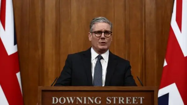 Keir Starmer wearing a black suit stands at a lecturn reading 'Downing Street' with two United Kingdom flags on either side.