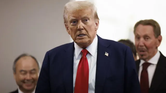 Donald Trump, wearing a blue suit and red tie, looks off to the left and pulls a facial expression; there are a group of men standing behind him.