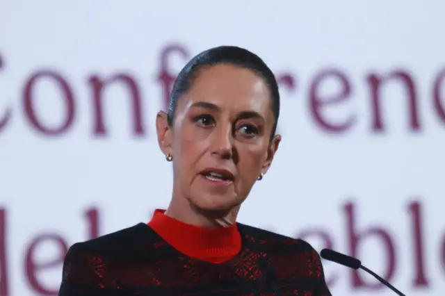 Sheinbaum with hair pulled back in red and black top in front of red letters on white screen