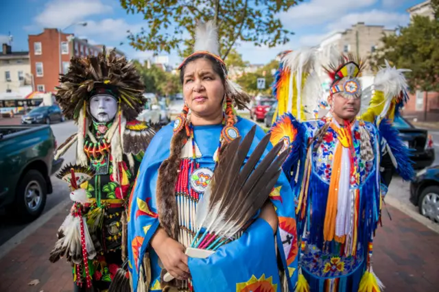 Three people on a city street in very colourful dress, including robes, feathers and headdresses