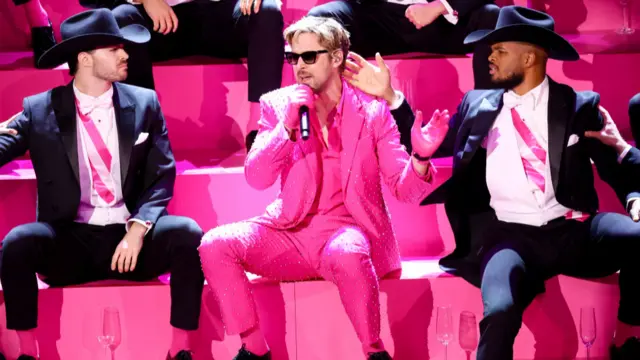 Ryan Gosling performs at the 2024 Oscars ceremony wearing a pink suit and shirt, with black shoes and sunglasses, surrounded by dancers wearing black and white suits with pink ties. Ryan is sitting on pink stairs holding a microphone.