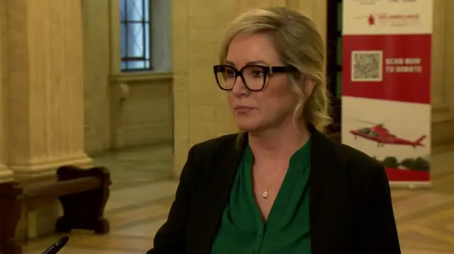 Michelle O'Neill, with shoulder-length blonde hair, a dark jacket and a green shirt standing in Stormont's Great Hall. In the background, we can see marble pillars and a window.