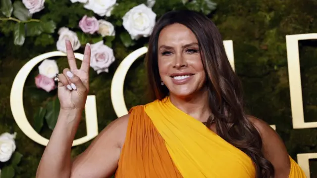 Close up shot of Karla Sofía Gascón wearing an orange and yellow dress. She is raising her hand into a peace sign.