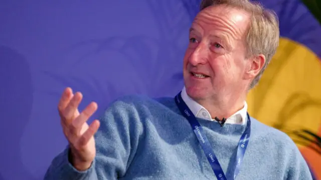 Sir Alex Younger, gesturing with his right hand as he speaks, shot from chest up, wearing a blue jumper