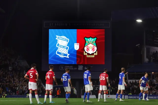 Birmingham v Wrexham, players lined up, probably at a set-piece, with the scoreboard in the background showing the logos of both clubs in a split-screen