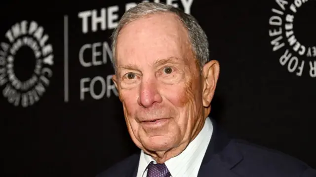 Michael Bloomberg wearing a dark suit in front of a black partition