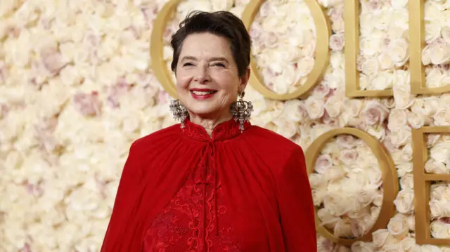 Isabella Rossellini wears a red dress with a thin red cape tied round her neck. She is smiling. She has short brown hair.