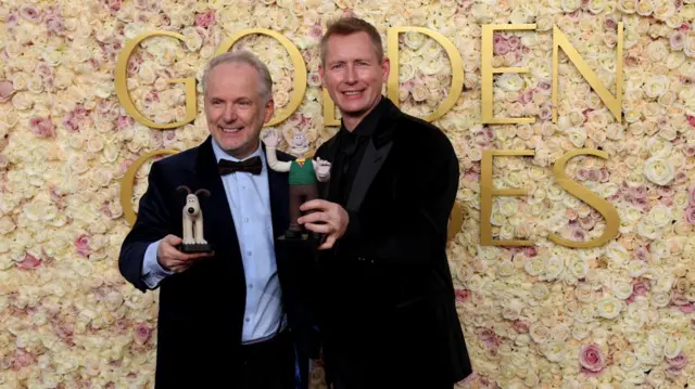 Nick Park and Merlin Crossingham wear suits in front of a Golden Globes sign holding Wallace and Gromit figurines