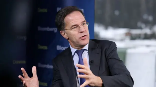 Mark Rutte wearing a blue suit, his hands are outstretched in front of him. There is a window and a blue banner in the background.