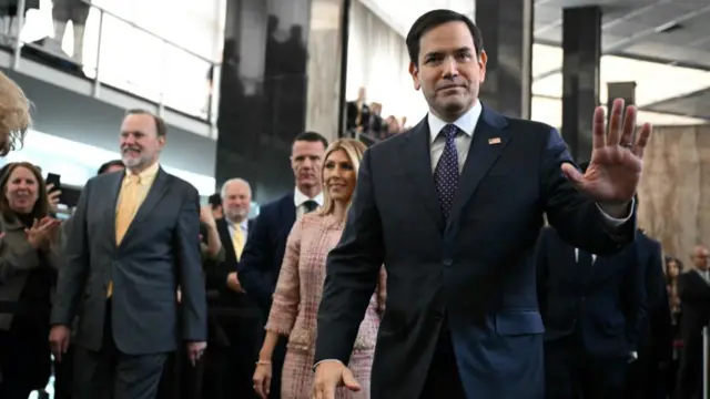 Marco Rubio walks in front of several people wearing a suit and a US pin on his lapel. He holds on hand up