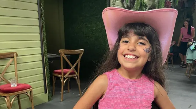 Alice da Silva Aguiar wears a pink top and a pink hat with glitter on her cheeks. She smiles and looks at the camera