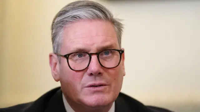 Close up of Keir Starmer wearing black suit, black rimmed glasses while sitting down. Behind him is a cream yellow wall