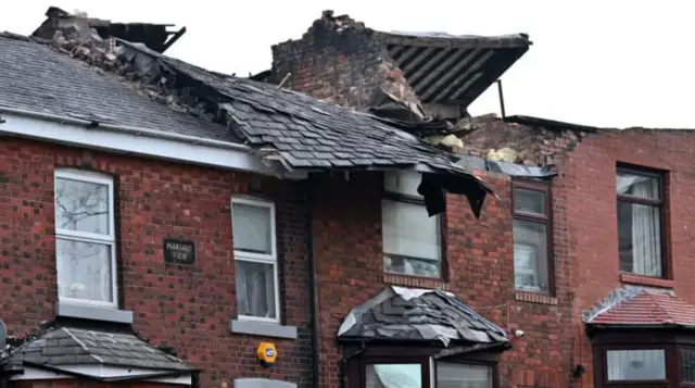 A row of red brick houses with black rooves with one of the rooves blown off