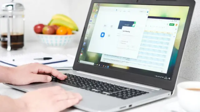 A laptop open, there is hands moving the mouse, a bowl of fruit is behind in the background