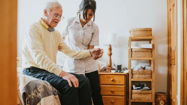 A care worker helps an elderly man get up from a sitting position on his bed