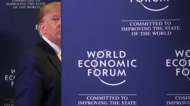 Donald Trump walking behind a sign. The sigin is dark blue and says "world economic forum committed to improving the state of the world" in white