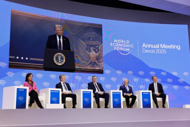 Video projection of Trump in dark suit above line of executives sitting in white chairs