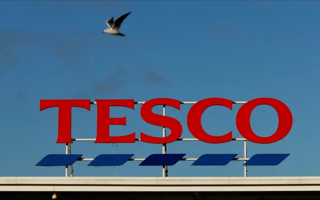 A Tesco supermarket sign on top of a building. The Tesco letters are coloured red, with blue underlining