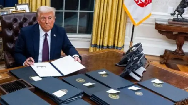 President Trump sat behind a brown desk. There are several piles of leather binders in front of him. The wall behind him is white and yellow curtains are on either side of the window