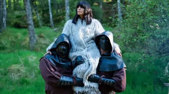 Claudia Winkleman wearing a fur coat and being carried by two masked men