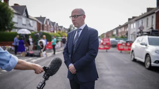 Patrick Hurley, Southport’s MP, wearing blue suit on road where attack happened, microphone in shot and cars in background