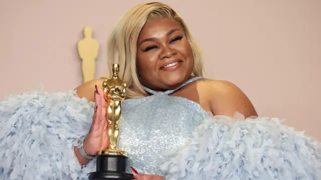 Da'Vine Joy Randolph holds up an Oscar while smiling for the camera. She has blonde hair and is wearing a blue-lilac dress with voluminous, feathery sleeves.