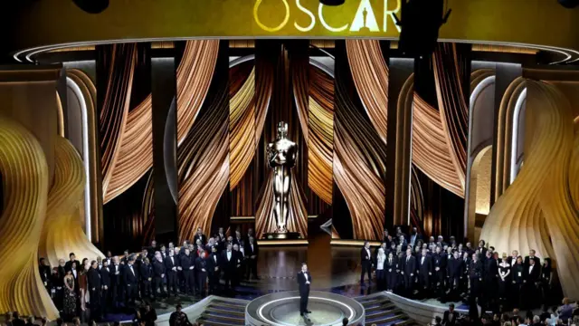 Jimmy Kimmel stands on a stage in front of an audience. Behind him is a large screen with golden curtains and a massive Oscars trophy in the centre. Kimmel is wearing a tuxedo and has brown hair.